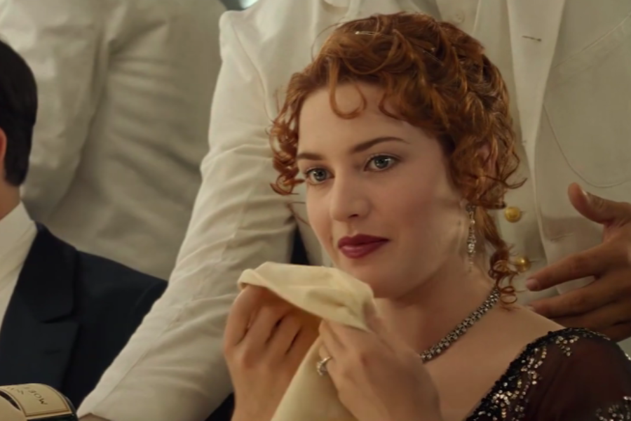 Close-up of Rose from Titanic smiling and holding a napkin, wearing an elegant gown and diamond necklace while dining.