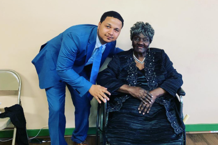 A man in a blue suit poses beside an elegantly dressed elderly woman in a black outfit seated in a wheelchair.