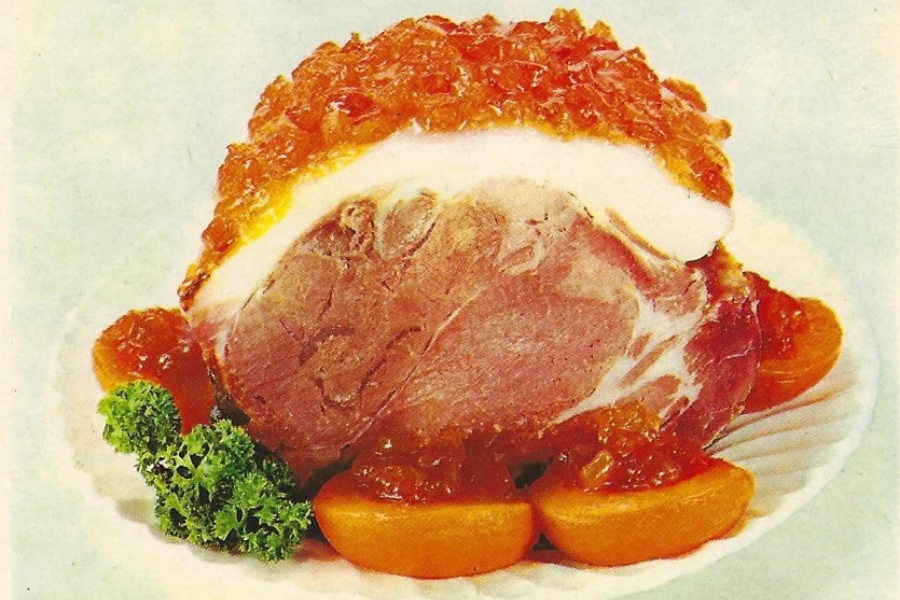 A large glazed ham topped with a chunky golden marmalade, surrounded by halved canned peaches and parsley on a white plate.