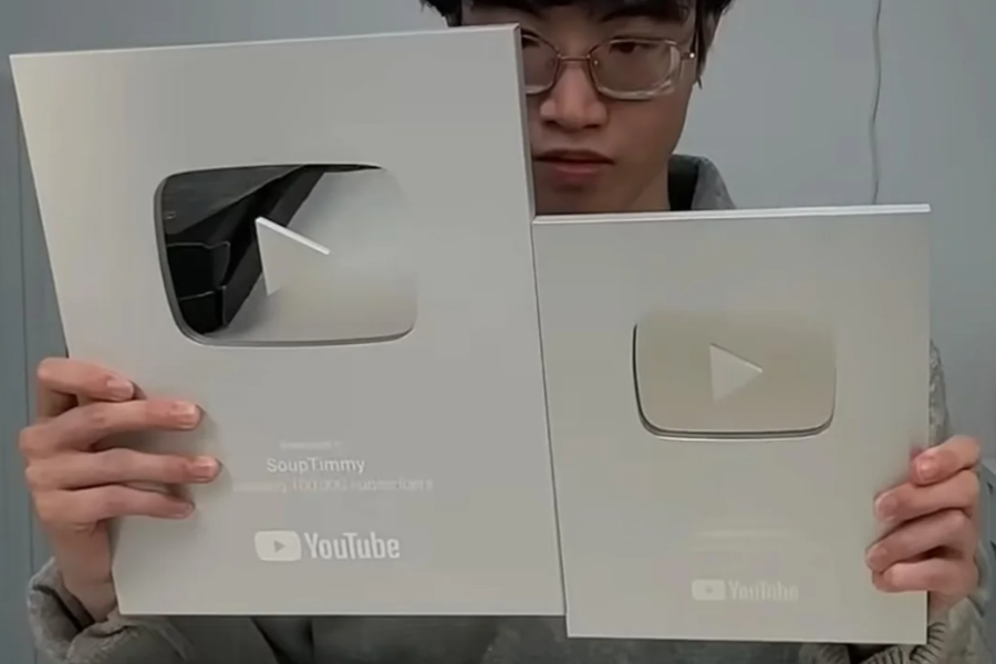 A person holding two YouTube silver play buttons, showing the old larger version in the left hand and the newer, smaller one in the right.