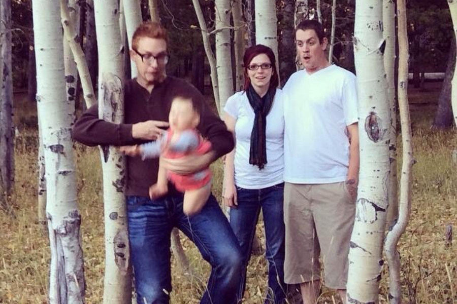 A man accidentally blurs his baby mid-motion while posing with two others among tree trunks; their surprised faces say it all.