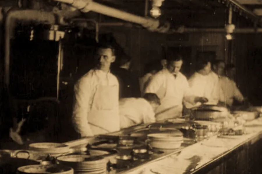 Sepia-toned photo of Titanic kitchen staff at work, with chefs preparing food at long counters under exposed piping.