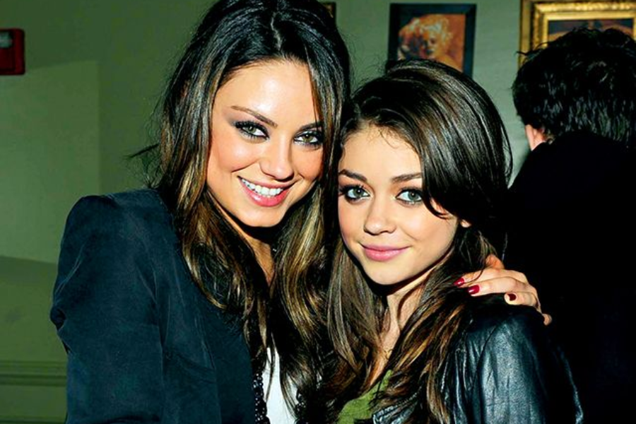 Two women with long dark brown hair smiling and posing closely together indoors. The woman on the left wears a dark blazer and has her arm around the woman on the right, who wears a black jacket and olive top.