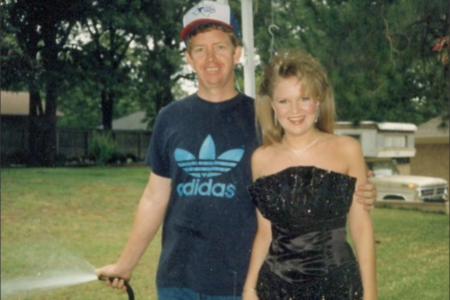 A man in a casual outfit waters the lawn while posing next to a young woman dressed for prom, creating a wildly mismatched moment.