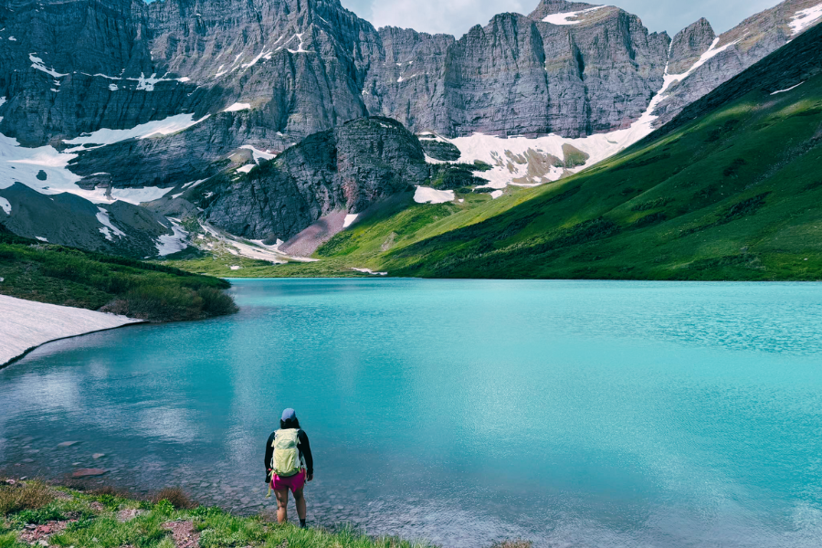A hiker with a backpack stands at the edge of a turquoise alpine lake surrounded by snow-dusted cliffs and lush green slopes in Glacier National Park.