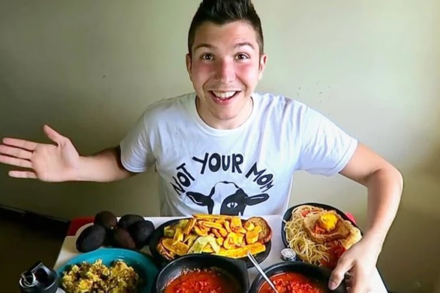 Smiling man in a “Not Your Mom” vegan-themed shirt sits in front of a table filled with a large spread of colorful food dishes including pasta, avocado, and beans.
