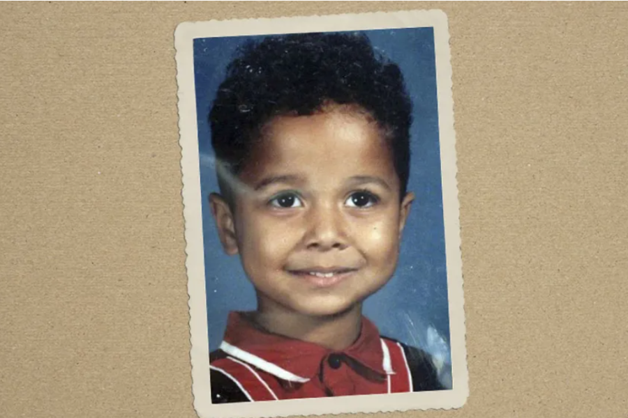 A childhood portrait of a smiling young boy with curly hair, wearing a red collared shirt, displayed on a scrapbook-like background.
