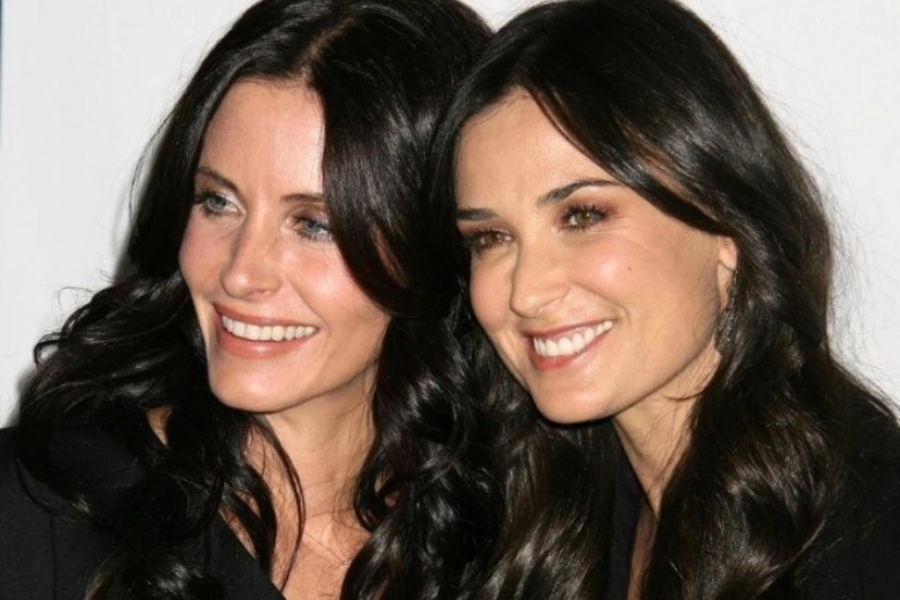 Two women with long wavy dark hair smiling and leaning close together for a photo. Both are wearing black outfits and posing against a light background.