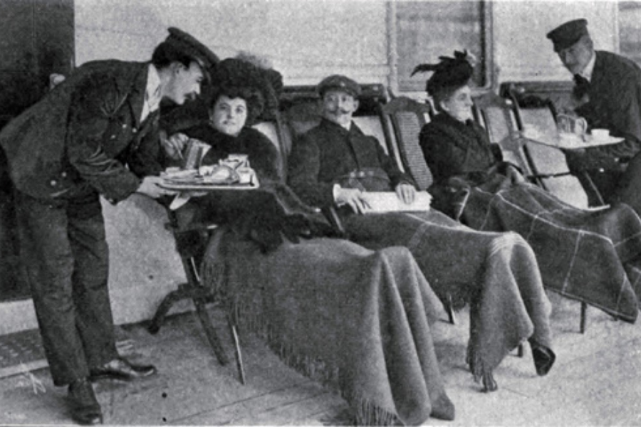 Vintage photo showing first-class passengers lounging under blankets as stewards serve them drinks on the Titanic’s promenade deck.