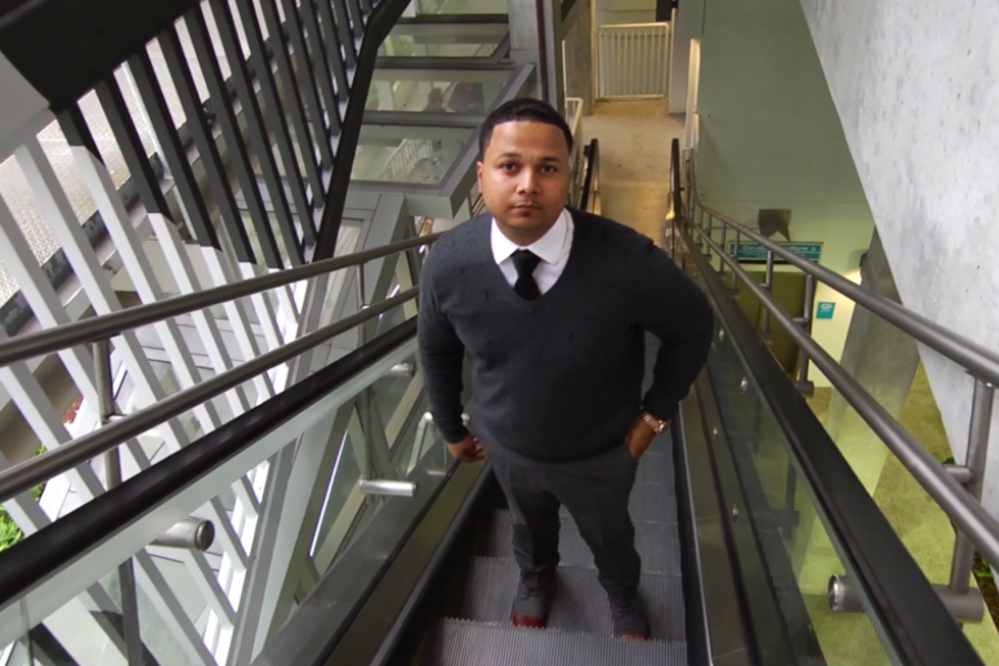 A man in business attire rides an escalator in a modern, industrial-looking building, gazing upward at the camera.