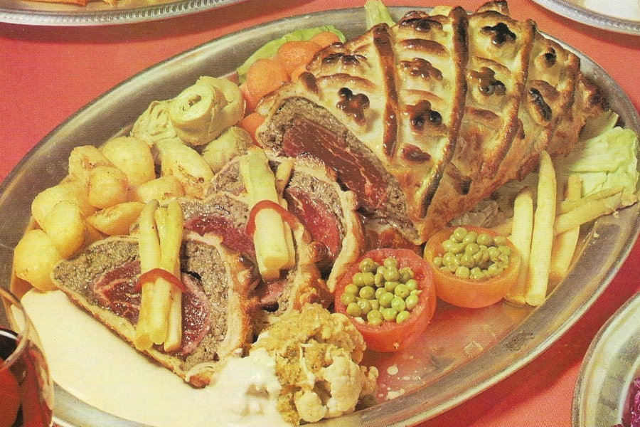 A large beef Wellington-style roast with pastry lattice crust, sliced to reveal a rare red center and topped with bundles of white asparagus.