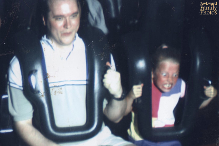 A father and daughter ride a theme park roller coaster; the dad looks relaxed while the girl appears utterly terrified, eyes wide and mouth clenched.