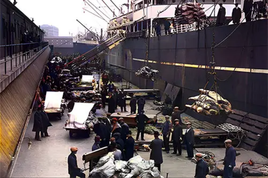 Colorized photo of Titanic’s provisioning with large crates of food being loaded onto the ship by dock workers using cranes and carts.