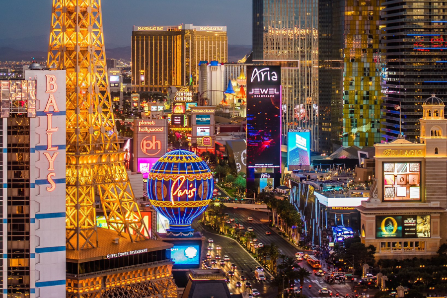 A nighttime view of the Las Vegas Strip glows with colorful neon signs, including replicas of the Eiffel Tower and Paris balloon among luxury hotels and casinos.