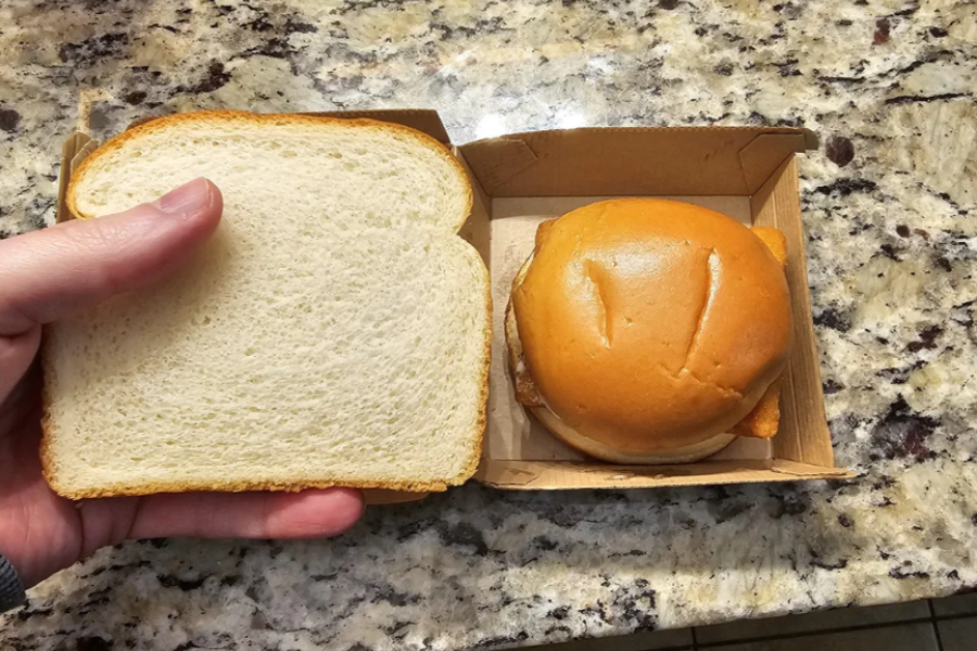 A slice of plain sandwich bread held next to a small fast food chicken sandwich, highlighting the sandwich's small size in comparison.