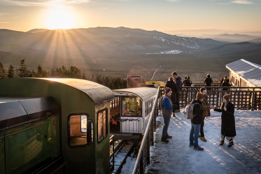 Visitors gather at a snowy mountain summit at sunrise beside the Mount Washington Cog Railway train, overlooking distant ridges and valleys.