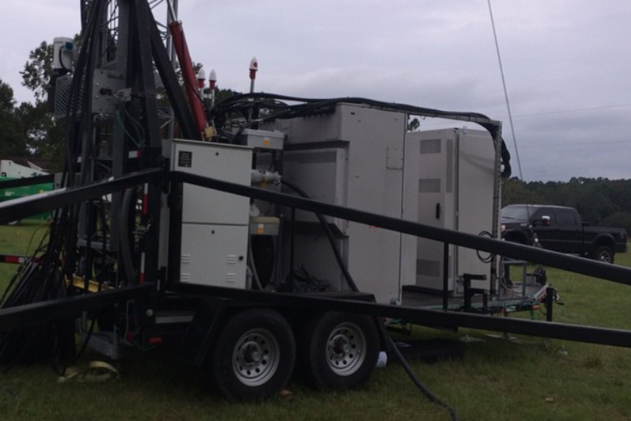 Side view of a mobile communications trailer with multiple cabinets and cables, set up in a grassy field with vehicles nearby.