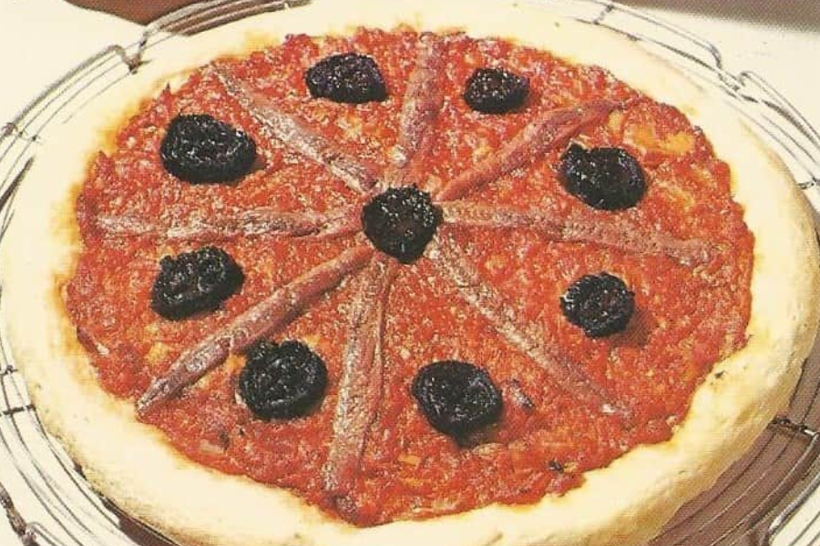 A pizza with a thick crust, topped with tomato sauce, anchovy fillets arranged in a radial pattern, and large black olives placed between the fillets.