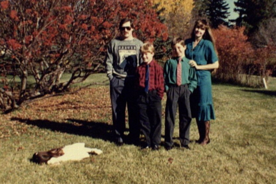A family poses cheerfully outdoors while a kid lies motionless on the grass in the foreground, stealing the scene unintentionally.