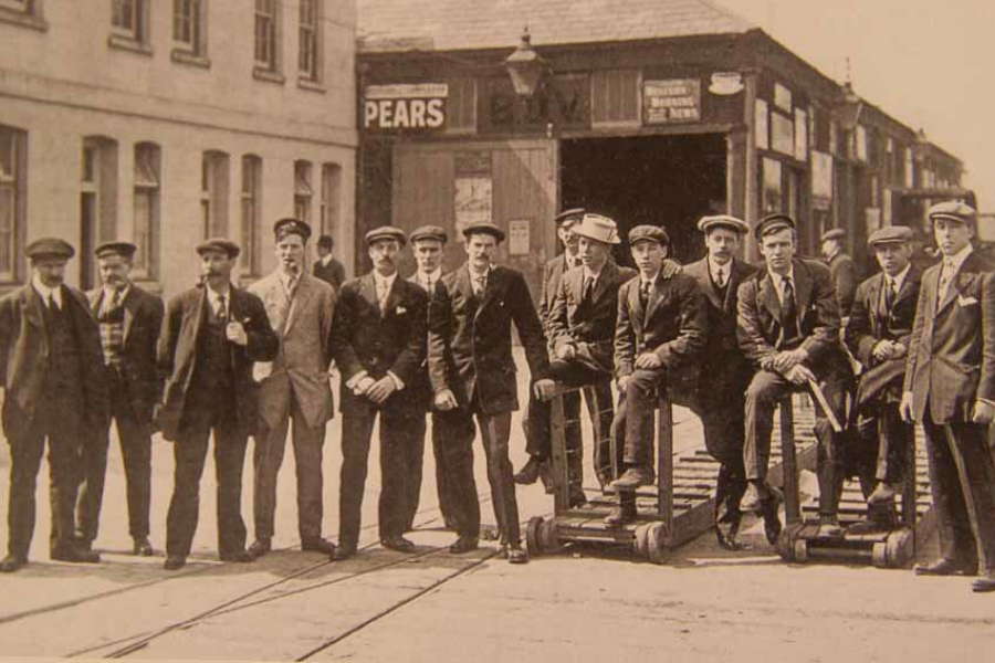 Group of young male dock workers posing with wooden carts outside a shipping warehouse in the early 1900s.