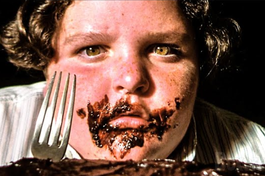 Close-up of a child with chocolate smeared around their mouth and a fork held upright beside a large chocolate cake.
