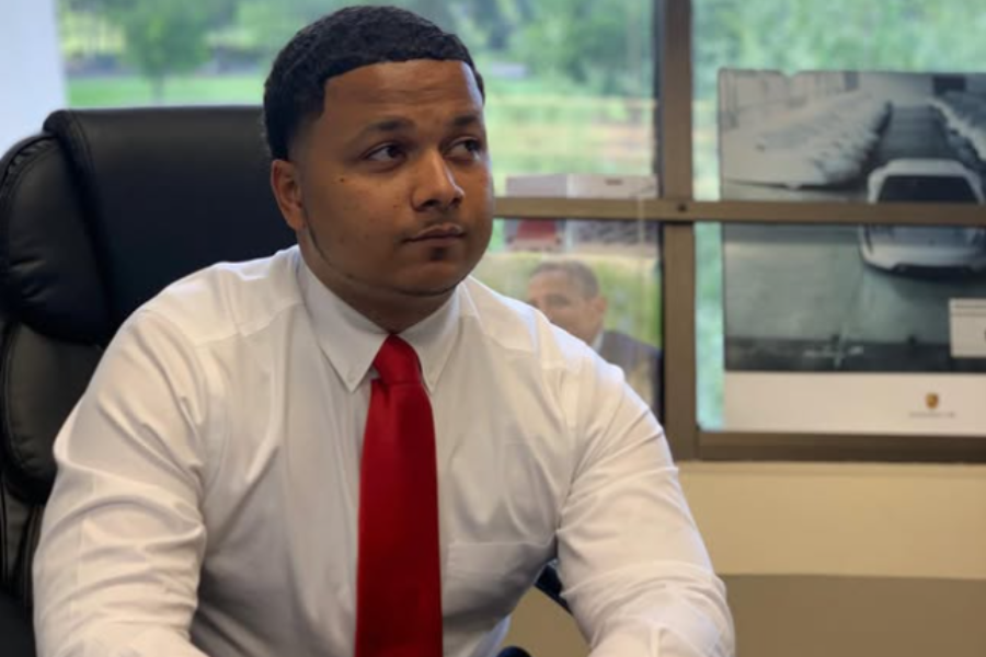 A man in a white shirt and red tie sits in a black office chair, looking to the side, with car posters and a window in the background.
