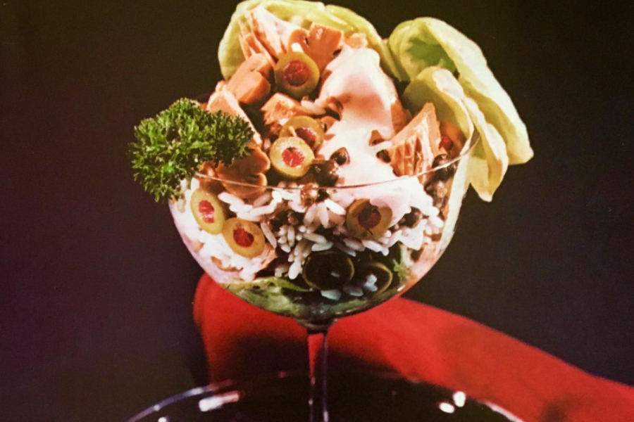 A layered salad served in a cocktail glass with green olives, rice, lettuce, canned vegetables, and pink dressing, garnished with a sprig of parsley.