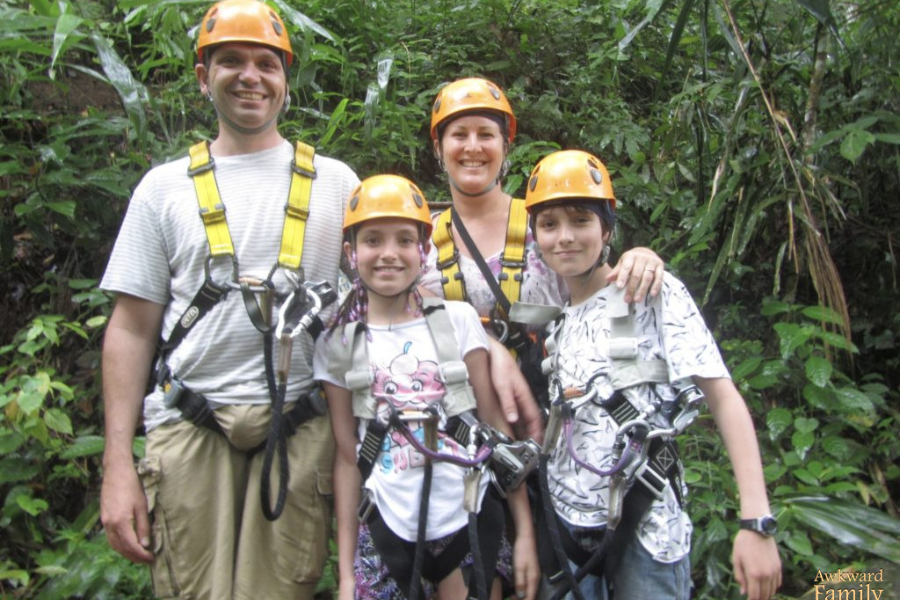 A family in zipline harnesses and helmets smiles for a photo in the jungle, though their gear slightly overwhelms their outfits.