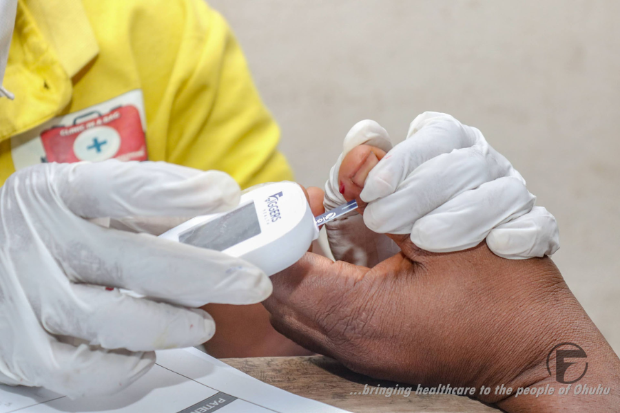 Gloved hands use a Figgers blood glucose meter to test a person’s finger during a healthcare check-up, with the tagline “bringing healthcare to the people of Ohuhu” visible.