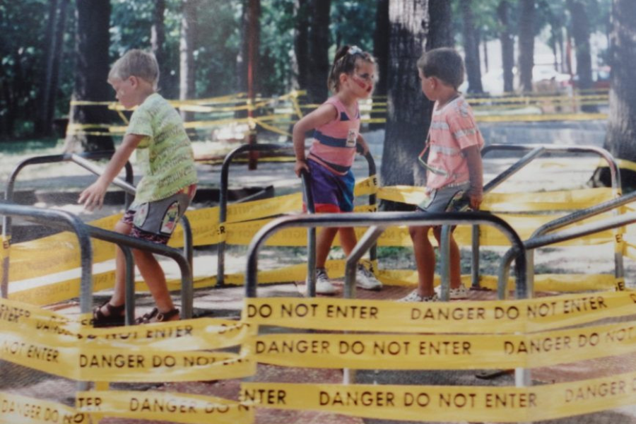 Three kids play on a merry-go-round completely surrounded by caution tape that reads “DANGER DO NOT ENTER.”