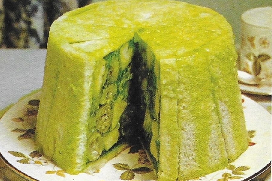 tall dome-shaped loaf lined with white bread slices, filled with green gelatin and layered vegetables, resembling a retro savory charlotte.