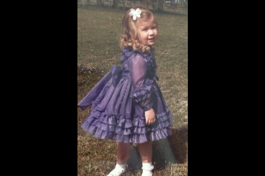 A little girl in a frilly purple dress stands in a yard, smiling sweetly while accidentally appearing to pee on a tree due to an unfortunate angle.