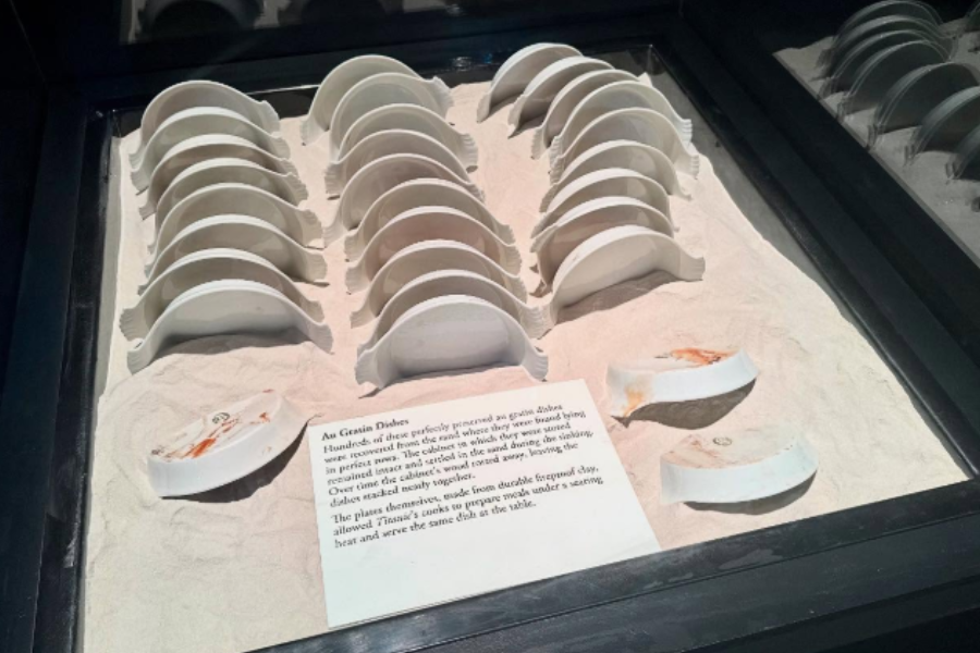 Museum display of intact and broken white au gratin dishes from the Titanic, arranged in sand with a descriptive placard.