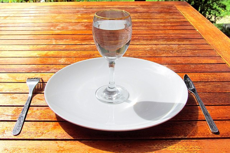 Empty white plate with a glass of water placed on it, set on a wooden table with a knife and fork on either side.