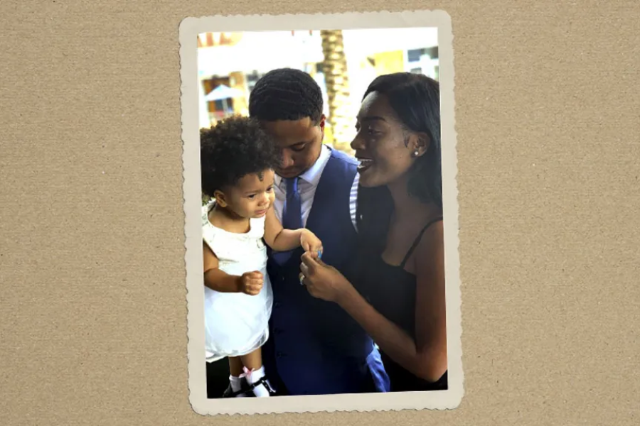 A candid family moment showing a man in a blue suit and a woman in a black dress engaging playfully with a toddler in a white outfit.