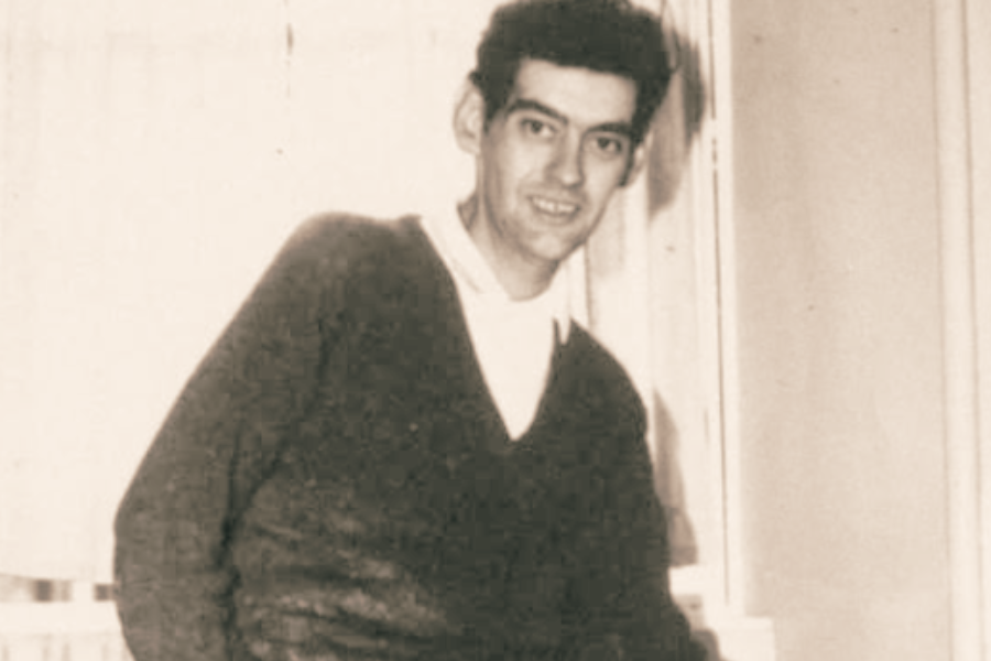 Sepia-toned photo of a slim man in a sweater leaning slightly forward and smiling toward the camera in a softly lit room.