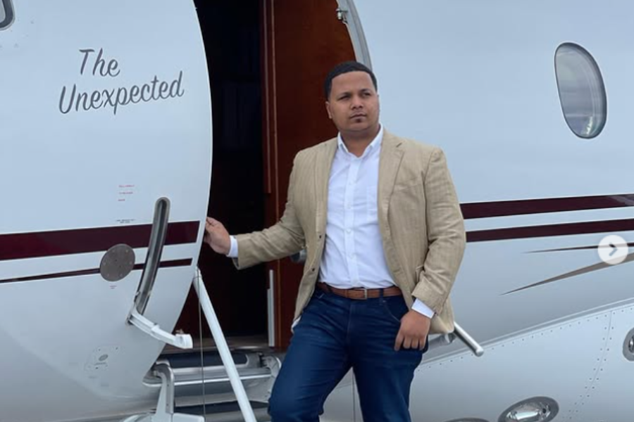 A man in a tan blazer steps confidently out of a private jet labeled “The Unexpected,” holding the door rail.