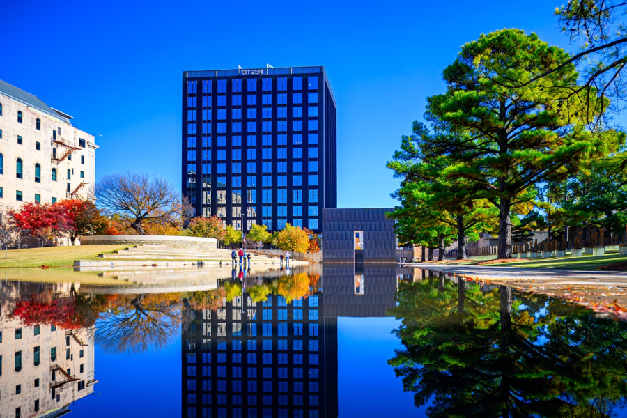 A calm reflecting pool mirrors the skyline and trees at the Oklahoma City National Memorial, with visitors walking quietly near the Field of Empty Chairs.