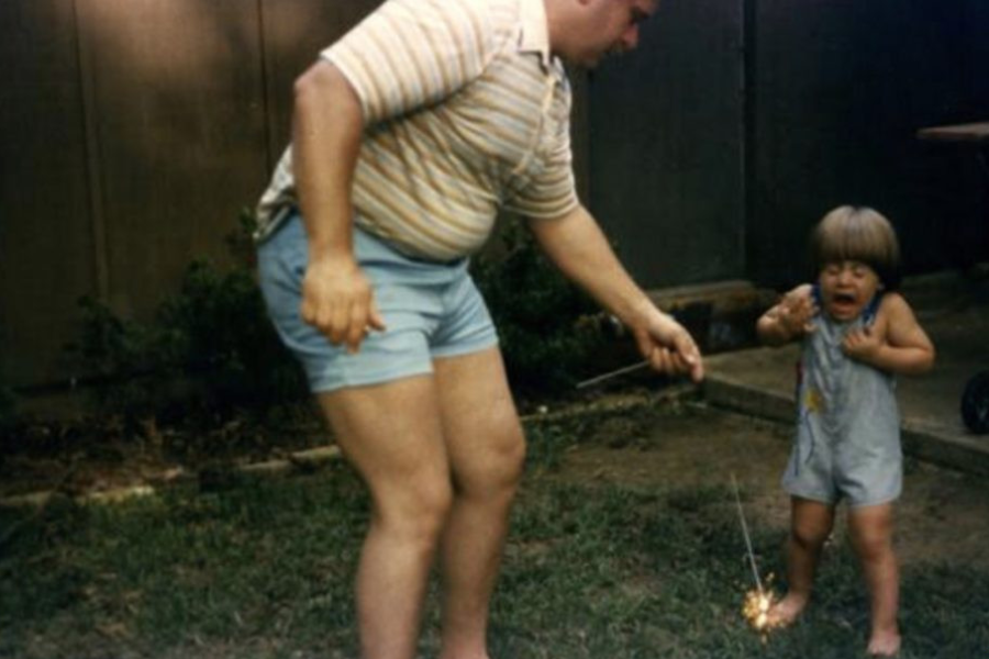 A man lights a sparkler while a nearby child recoils in fear, mouth open in mid-scream and arms drawn back.