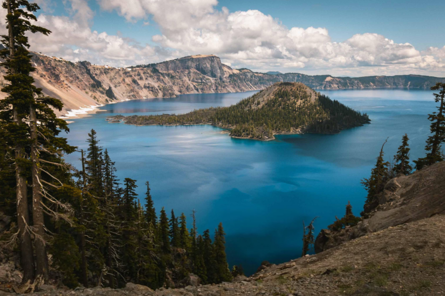 Wizard Island rises from the deep blue waters of Crater Lake, surrounded by steep cliffs and evergreen trees under a partly cloudy sky.