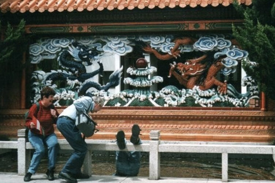 A tourist appears to have fallen backward over a stone bench, feet in the air, while two companions react mid-laugh and mid-rescue.