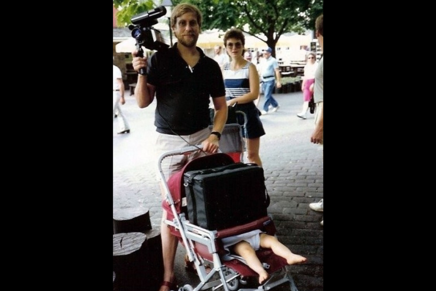 A man proudly holds a large camcorder while pushing a stroller completely taken over by a bulky black case, with a baby’s legs awkwardly poking out.
