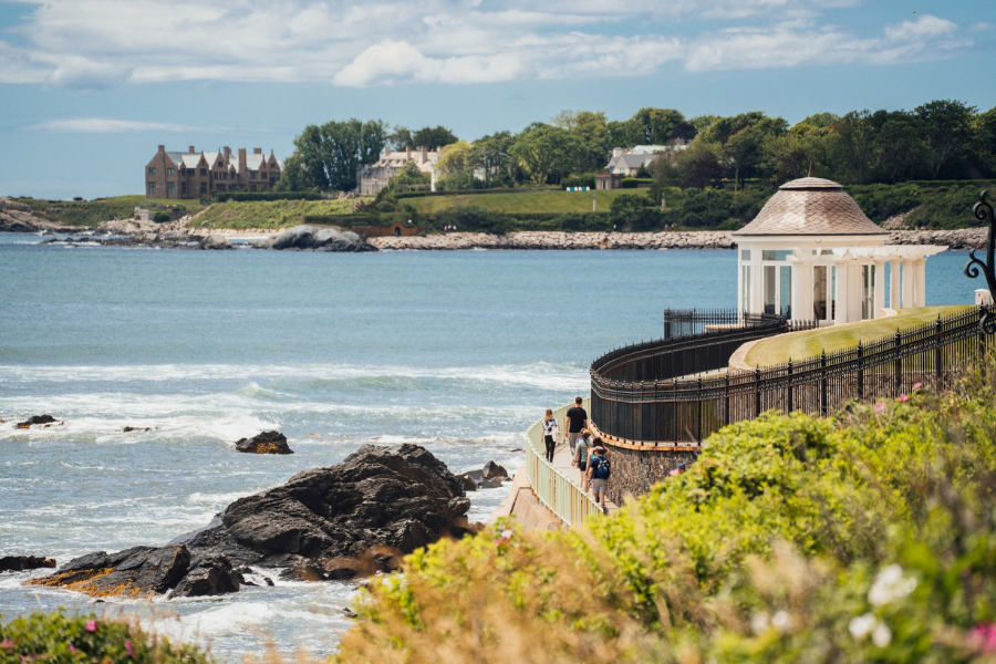 Tourists walk along the scenic Newport Cliff Walk with ocean waves crashing below and historic mansions overlooking the rocky coastline.