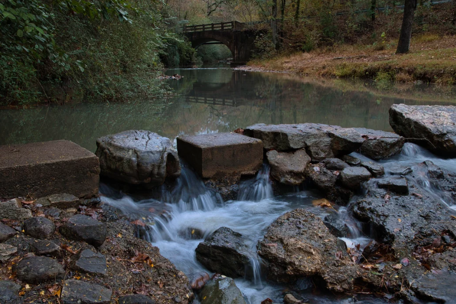A gentle stream flows over rocks and concrete blocks in a forested park area, with a small footbridge crossing the water in the background.
