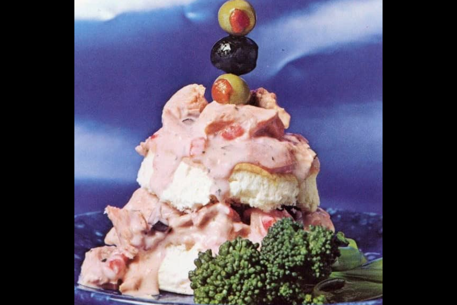 A layered dish of biscuit-like rounds topped with pink creamy sauce and diced vegetables, garnished with a skewer of green and black olives and served with broccoli.