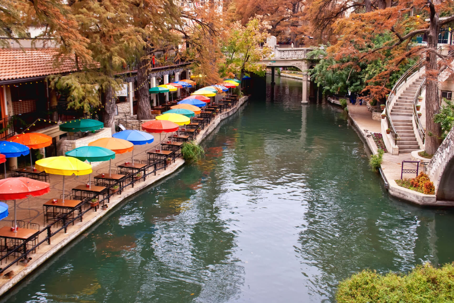 The San Antonio River Walk lined with colorful umbrellas and café tables beside the canal, surrounded by fall foliage and stone bridges.