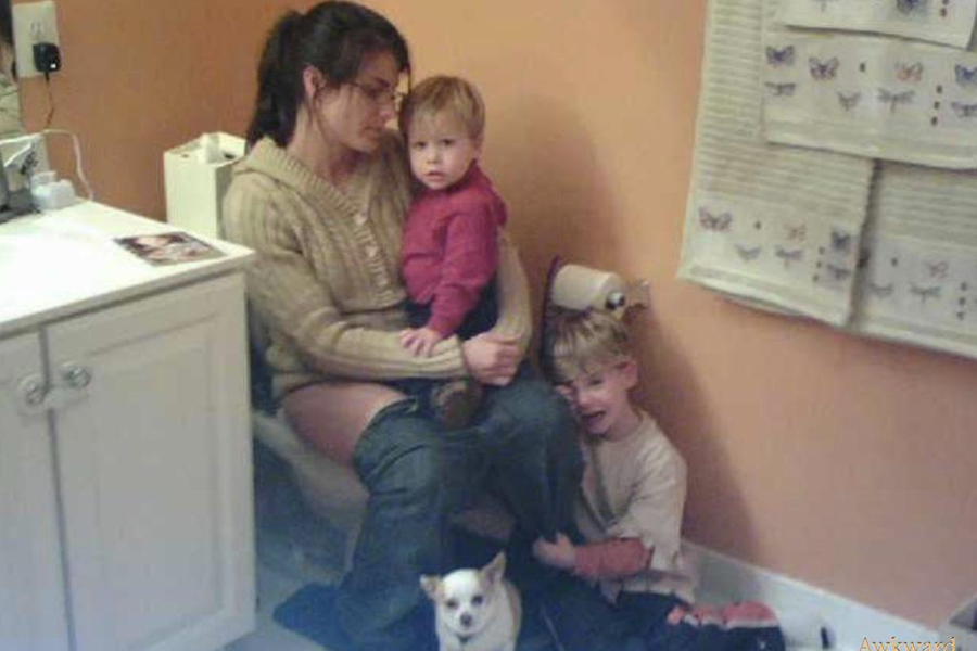 A mom sits on a toilet with her pants down while holding one child, another clings to her leg, and a chihuahua stands guard by her feet.