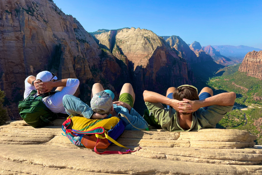 Three hikers lay back on a rocky ledge enjoying panoramic views of Zion Canyon’s towering sandstone cliffs under a clear blue sky.