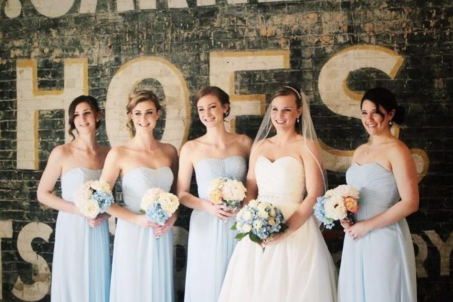 A bride stands with her bridesmaids in front of a brick wall that accidentally highlights the word “HOES” behind them.