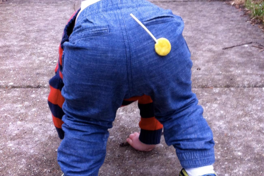 A baby crawling on a sidewalk has a bright yellow lollipop stuck squarely to their rear end, with the stick pointing up.